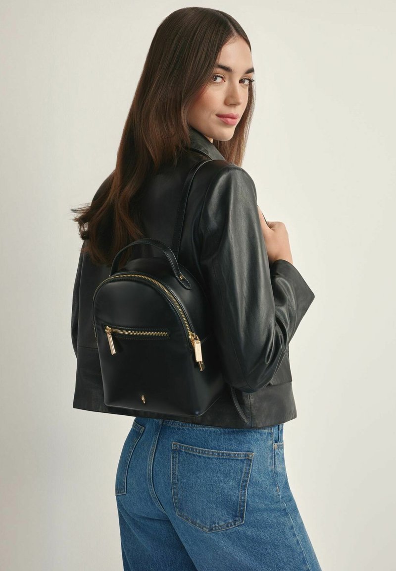 Kobieta o długich brązowych włosach, ubrana w czarną skórzaną kurtkę i niebieskie dżinsy, niesie mały czarny skórzany plecak z złotymi suwakami.