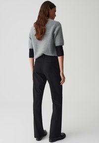 Vrouw met lang haar die een grijze fuzzy trui met korte mouwen over een zwart shirt met lange mouwen draagt, zwarte broek en zwarte schoenen, staande met haar rug naar ons gekeerd.