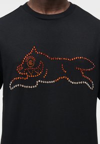 Μαύρο μπλουζάκι με σχέδιο γάτας διακοσμημένο με πορτοκαλί και διαφανείς στρας, που παρουσιάζει απαλή υφή και στρογγυλή λαιμόκοψη.