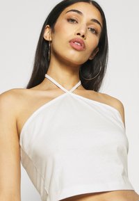 Top cropped branco feito de tecido suave, com um design de pescoço halter com alças cruzadas. Forma simples e ajustada com um acabamento limpo.