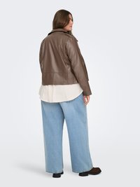 Frau trägt eine braune Lederjacke, lockere hellblaue Jeans, weißes Hemd und dunkle Schuhe, steht leicht nach links gewandt vor einem einfarbigen Hintergrund.