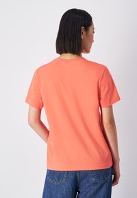 Koraalkleurig t-shirt met korte mouwen, gemaakt van zachte katoen. Klassiek ontwerp met een ronde hals en een relaxte fit, gezien vanuit de achterkant, gecombineerd met denim jeans.