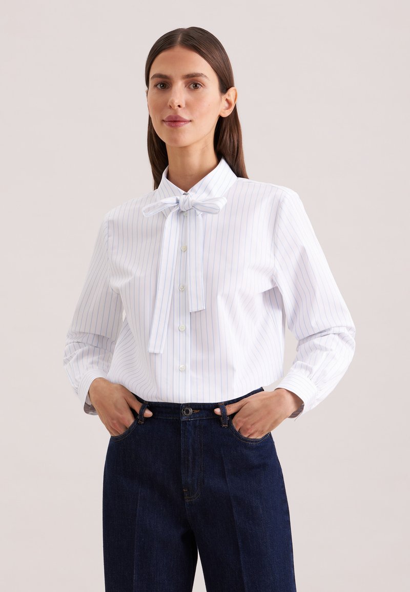 Femme portant un chemisier blanc à fines rayures verticales bleues et un nœud noué au col, rentré dans un jean taille haute bleu foncé, les mains dans les poches.