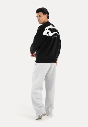 Uomo che indossa una felpa nera con grafica bianca sulla schiena e pantaloni della tuta grigio chiaro, in piedi che guarda oltre la spalla.