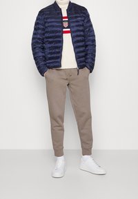 Marinblå puffjacka med quiltat mönster, krämfärgad stickad tröja med röda och blå ränder, beige mjukisbyxor och vita sneakers.