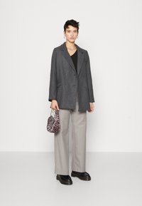 Grau doppelt gesteppten Mantel, schwarzes Oberteil, hellgraue Nadelstreifenhose, schwarze klobige Schuhe und eine Tasche mit Leopardenmuster. Glatte Textur.