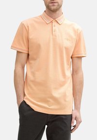 Polo shirt à manches courtes en coton doux pêche, avec un col à rayures blanches et une patte de boutonnage à trois boutons. Détail de logo subtil.
