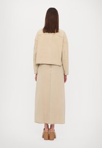 Giacca di denim beige con un design corto, abbinata a una lunga gonna beige. Entrambi hanno orli grezzi. Le scarpe con tacco alto marroni completano l'outfit.
