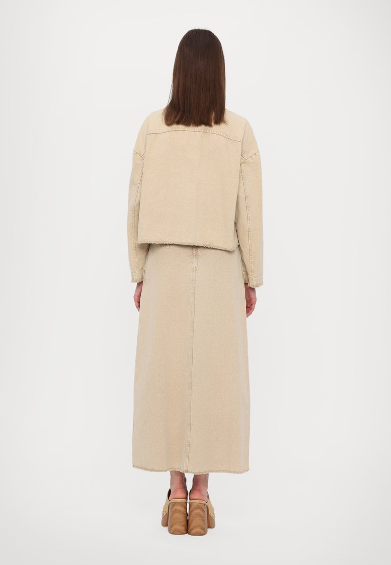 Beige denimjack met een cropped ontwerp, gecombineerd met een lange beige rok. Beide hebben onafgebroken zomen. Bruine platform schoenen maken de outfit af.