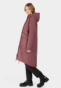 Long burgundy waterproof coat with a hood, front pockets, and a waist tie, paired with black ankle boots and dark jeans. Smooth texture.