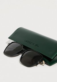 Čierne obdĺžnikové slnečné okuliare s lesklým povrchom, položené vedľa tmavozeleného koženého puzdra s vyrazeným nápisom "LACOSTE".