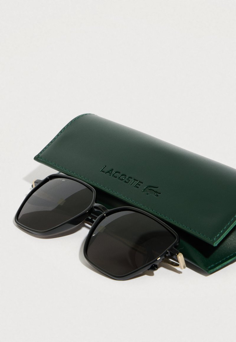 Sorte rektangulære solbriller med en blank finish, placeret ved siden af et mørkegrønt læderetui præget med "LACOSTE."