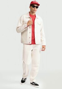 Λευκό τζιν μπουφάν πάνω από κόκκινο πουλόβερ με πλέξη καλώδιο, σε συνδυασμό με λευκό παντελόνι και μαύρα αθλητικά παπούτσια, ολοκληρωμένο με κόκκινο καπέλο και γυαλιά ηλίου.