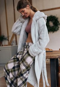 Lichtblauwe, zachte badjas met capuchon, gedragen over een mauve bralette en geruite pyjamabroek in groen, zwart en wit. Zachte texturen overheersen.