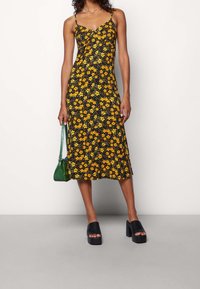 Svart klänning med gul blommönster, spagettiband, knälång, med en sidöppning. Bärs med svarta platåsandaler och en grön handväska.