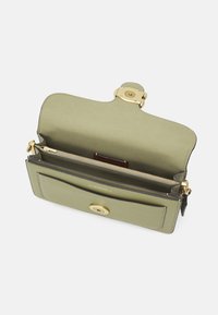 Jasnozielona skórzana torba na ramię z złotym zapięciem, otwarta, aby pokazać wewnętrzną kieszeń na zamek oraz zewnętrzną kieszeń wsuwaną z przodu.