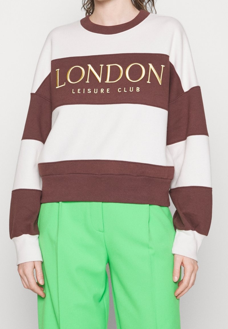 Sudadera color bloque marrón y crema con el texto dorado "LONDON LEISURE CLUB". Presenta un escote redondo y un ajuste relajado. Combinada con pantalones verdes.