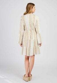 Weißes Kleid mit goldenen vertikalen Streifen, langen Glockenärmeln und einer gerafften Taille. Verfügt über gestufte Rüschen und einen strukturierten Stoff.