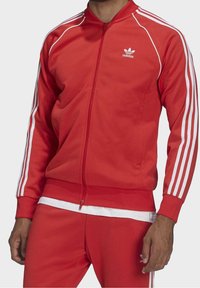 Red athletic tracksuit made of smooth fabric, featuring white three-stripe accents on sleeves and pants, with a zippered front and classic logo.