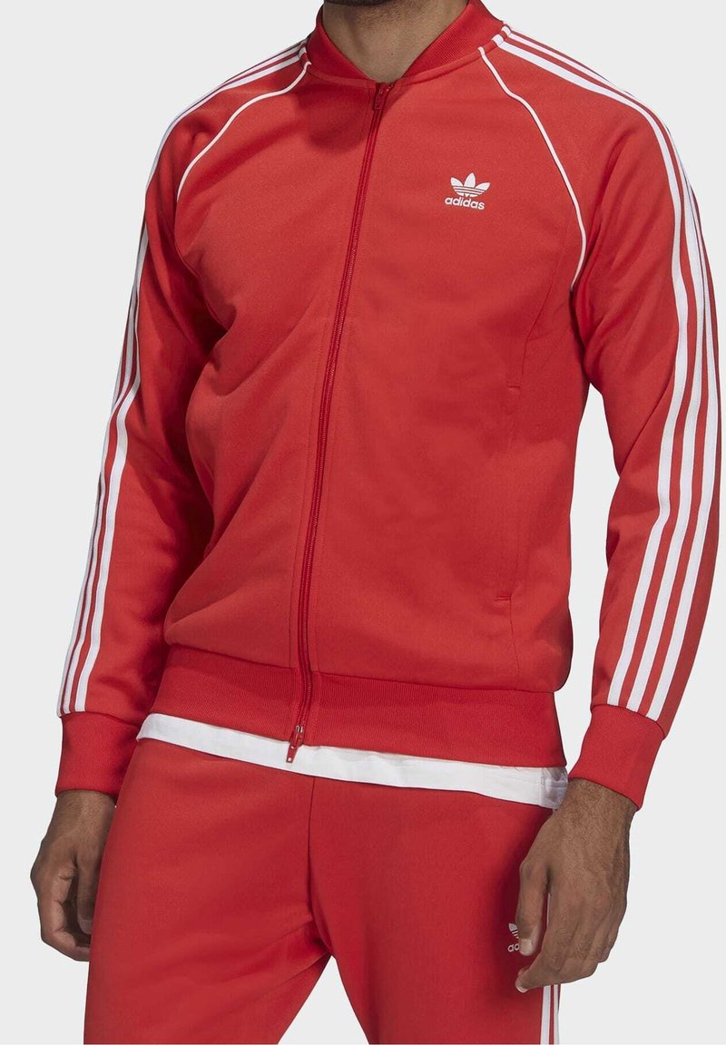 Red athletic tracksuit made of smooth fabric, featuring white three-stripe accents on sleeves and pants, with a zippered front and classic logo.