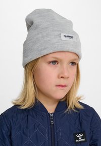 Grå stickad mössa med uppvikt kant, med en rektangulär etikett som säger "hummel sportswear". Barn som har på sig en marinblå vadderad bomberjacka.