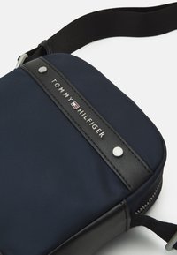 Borsa blu navy in nylon con un accento in pelle nera e dettagli in metallo argentato. Presenta il nome del marchio su una striscia orizzontale, con chiusura a zip.