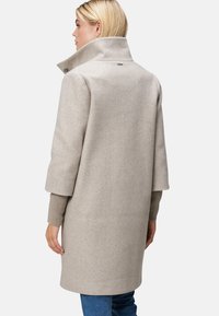 Manteau en mélange de laine beige avec un col montant, des manches amples et des accents en tricot côtelé aux poignets. Présente un logo subtil dans le dos.