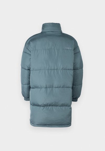 Duga puffer jakna u prigušenoj teal boji, s visokim ovratnikom, tkaninom s kvadratnim uzorkom i elastičnim manžetama. Suptilno brendiranje blizu vrata.