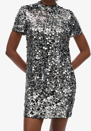 Kortærmet minikjole dækket af tæt pakkede sorte og sølvfarvede pailletter, båret af en person med armene bag ryggen.