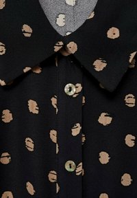 Blouse noire avec un col, ornée de pois beiges et agrémentée d'un détail de bouton clair.