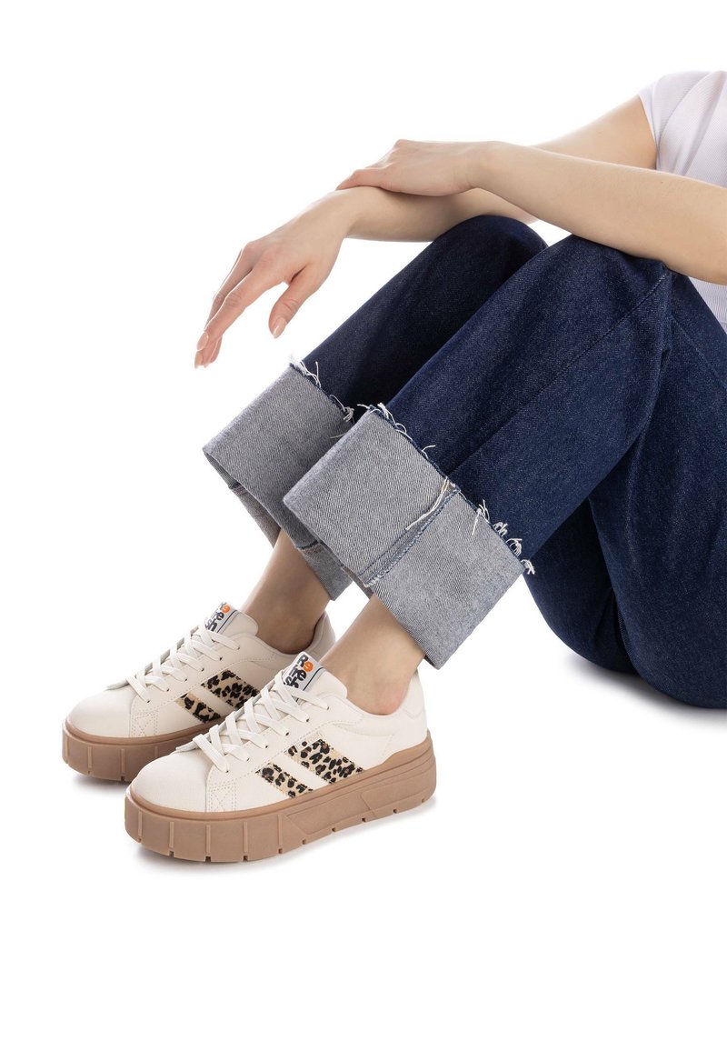 Zapatillas beige con acentos de leopardo negro, suela de goma texturizada y cordones planos, combinadas con pantalones recortados de mezclilla oscura.