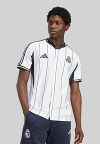 Jersey blanco a rayas con acentos en azul oscuro, presentado con una parte delantera botones, cuello y logo. Combinado con pantalones cortos azul oscuro.