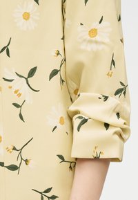 Κίτρινο ύφασμα με μανίκι μαζεμένο προς τα πάνω, διακοσμημένο με λευκά άνθη μαργαρίτας με κίτρινα κέντρα και πράσινα φύλλα πάνω σε υλικό με σχέδια.