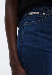 Blugi din denim albastru închis cu talie înaltă, având un patch Calvin Klein, nituri metalice și o textură netedă. Include buzunare frontale.
