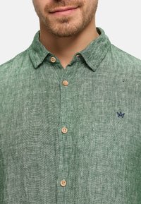 Nahaufnahme eines Mannes, der ein grünes, strukturiertes Hemd mit Knöpfen trägt, auf dessen Brust sich ein kleines gesticktes Logo befindet, und beigen Knöpfen.