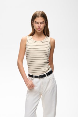 Jeune femme aux cheveux raides portant un haut beige sans manches avec de fines rayures horizontales et un pantalon blanc avec une ceinture noire.