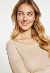 Beige neule, jossa on pehmeä ribattu rakenne, leveä kaula-aukko ja pitkät hihat. Mallilla on aaltoilevat vaaleat hiukset ja hienovarainen hymy.