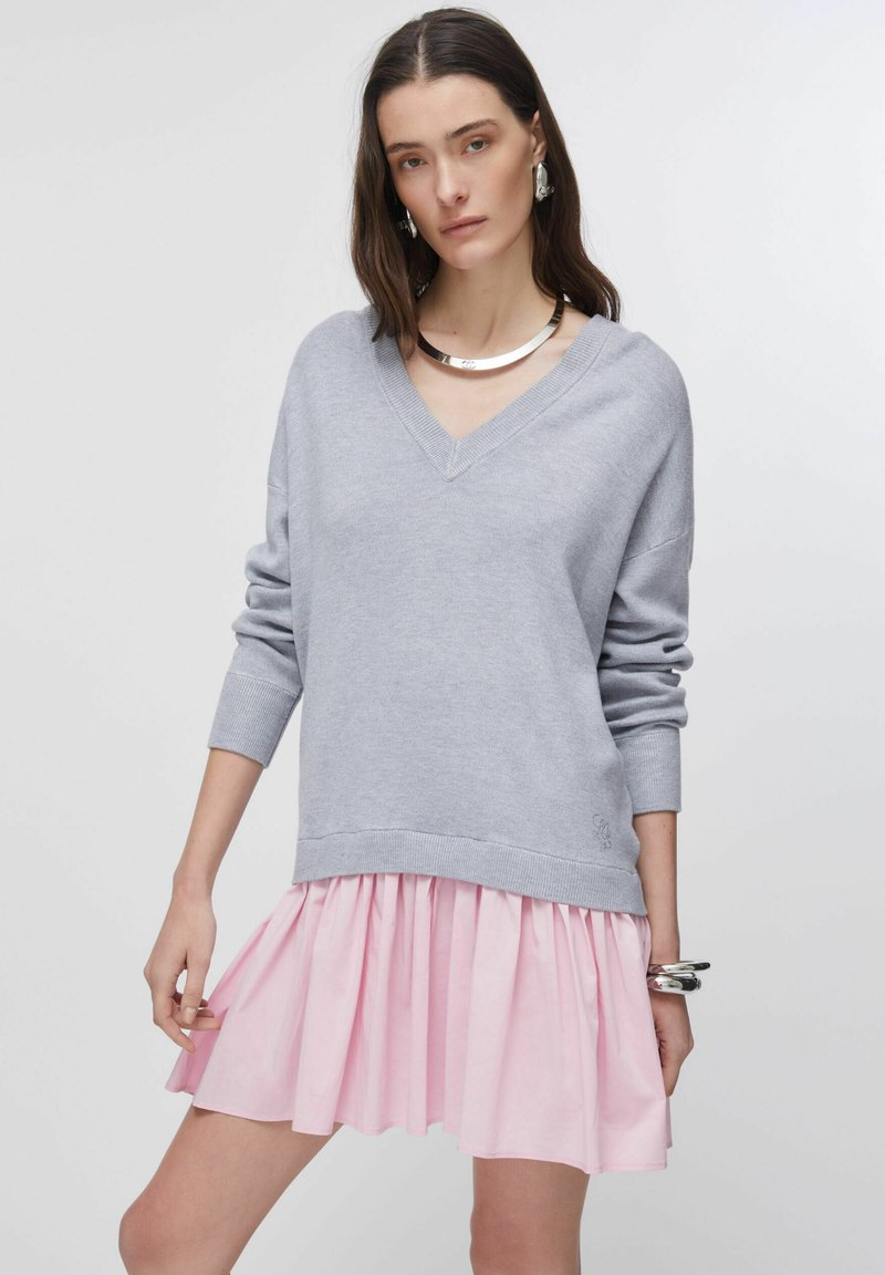 Pull gris à col en V avec manches longues, superposé sur une jupe plissée rose. Le pull est doté d'un ourlet et de poignets côtelés. Des bijoux argentés imposants viennent compléter le look.