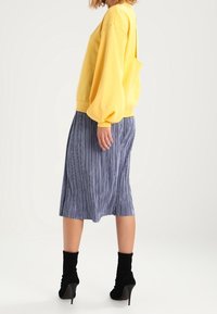 Felpa gialla oversize con spalle basse, abbinata a una gonna midi plissettata blu. Stivaletti neri completano l'outfit.