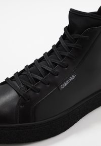 Černé vysoké tenisky vyrobené z kůže a textilu, s hladkým svrškem, strukturovanou podrážkou a černými tkaničkami. Logo na boku.