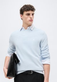 Νεαρός άνδρας που φοράει ανοιχτό μπλε πουλόβερ με υφή πάνω από ριγέ πουκάμισο κρατά τσάντα από μαύρο δερμάτινο χαρτοφύλακα, κοιτάζοντας προς το πλάι.