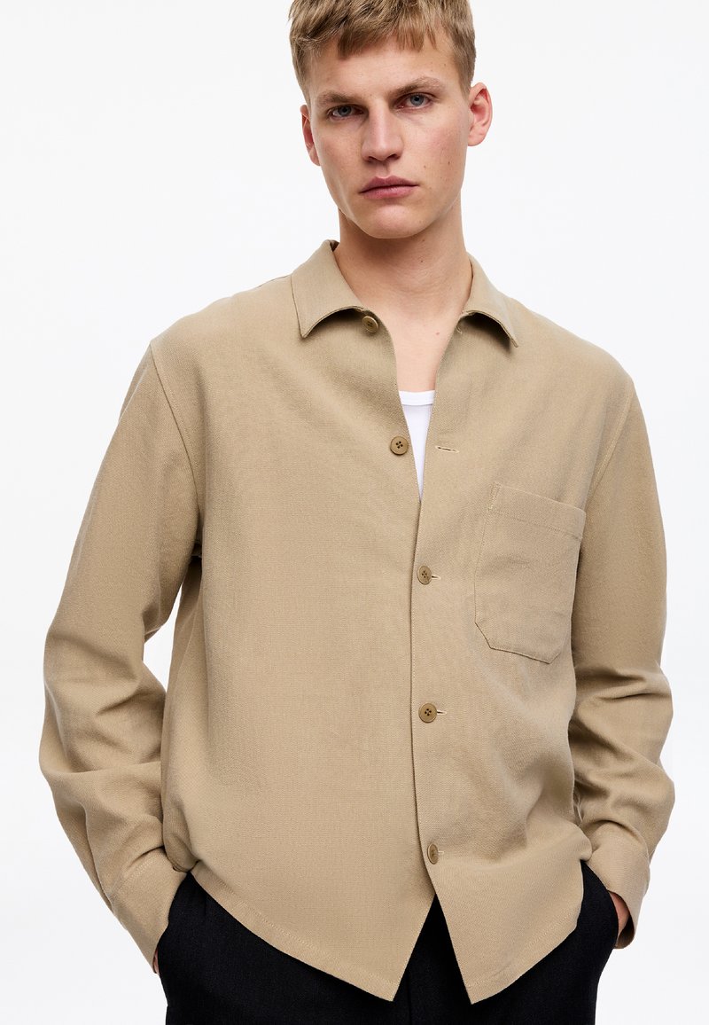 Jeune homme aux cheveux blonds courts portant une chemise beige boutonnée par-dessus un t-shirt blanc, mains dans les poches d'un pantalon sombre, fond neutre.