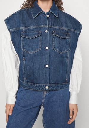 Blaue, ärmellose Jeansweste mit Knopfverschluss, getragen über einem weißen Langarmshirt, kombiniert mit passenden blauen Jeans.