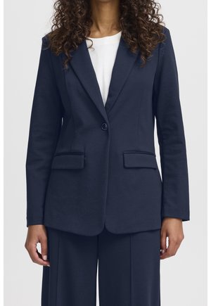 Frau trägt einen marineblauen Blazer mit einem Knopf und Pattentaschen, kombiniert mit passenden weit geschnittenen Hosen und einem weißen Top.