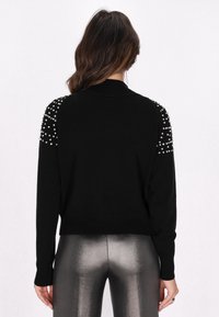 Maglione nero con un design attillato, caratterizzato da dettagli decorativi simili a perle sulle spalle e polsini a coste, abbinato a leggings argentati lucidi.