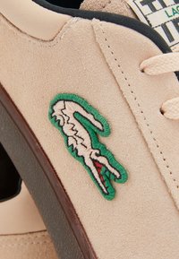 Zapatillas de ante beige con el logotipo de cocodrilo bordado en verde y blanco, cordones de color claro y suela negra texturizada.