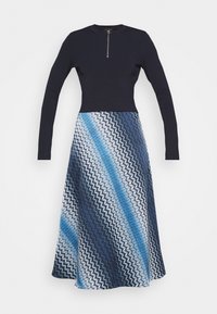 Långärmad marinblå klänning med dragkedja fram, figursydd överdel i enhetlig färg och utsvängd kjol med ett blått och vitt zick-zack-mönster.