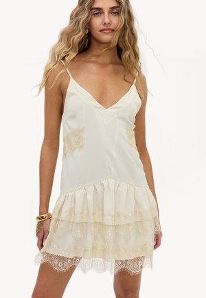 Femme aux longs cheveux blonds ondulés porte une robe nuisette crème sans manches avec des détails en dentelle et un bracelet en or à son poignet gauche.