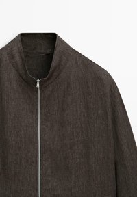 Brun jacka med dragkedja och hög krage, tillverkad av ett texturerat tyg, med en slimmad silhuett och minimal hårdvara.