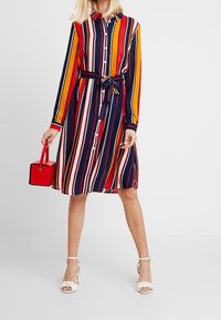 Robe à manches longues, boutonnée, avec des rayures verticales en bleu marine, rouge, jaune et blanc. Comprend une ceinture et un sac à main carré rouge. Sandales blanches.